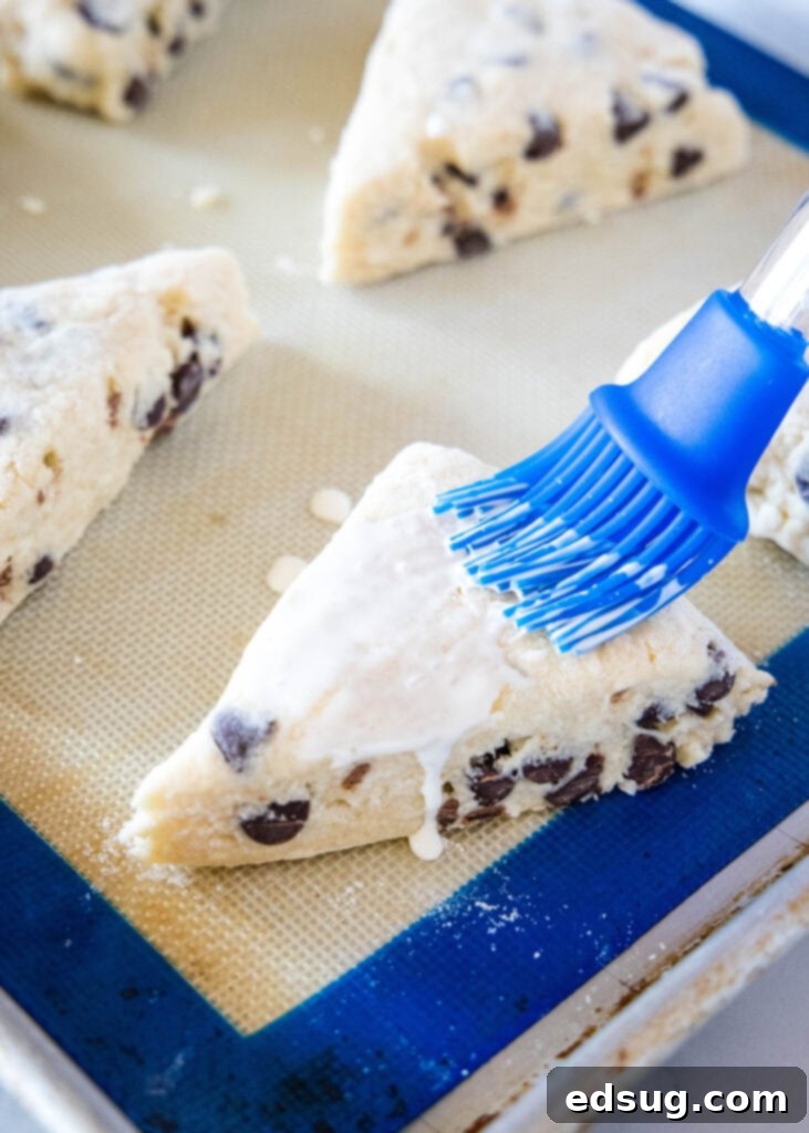A hand brushing heavy cream onto scone wedges on a baking sheet before baking, with coarse sugar visible.