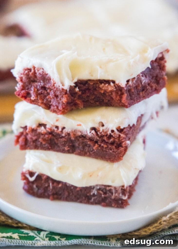 Stacked fudgy red velvet brownies on a white plate with one showing a bite mark, topped with fluffy cream cheese frosting.