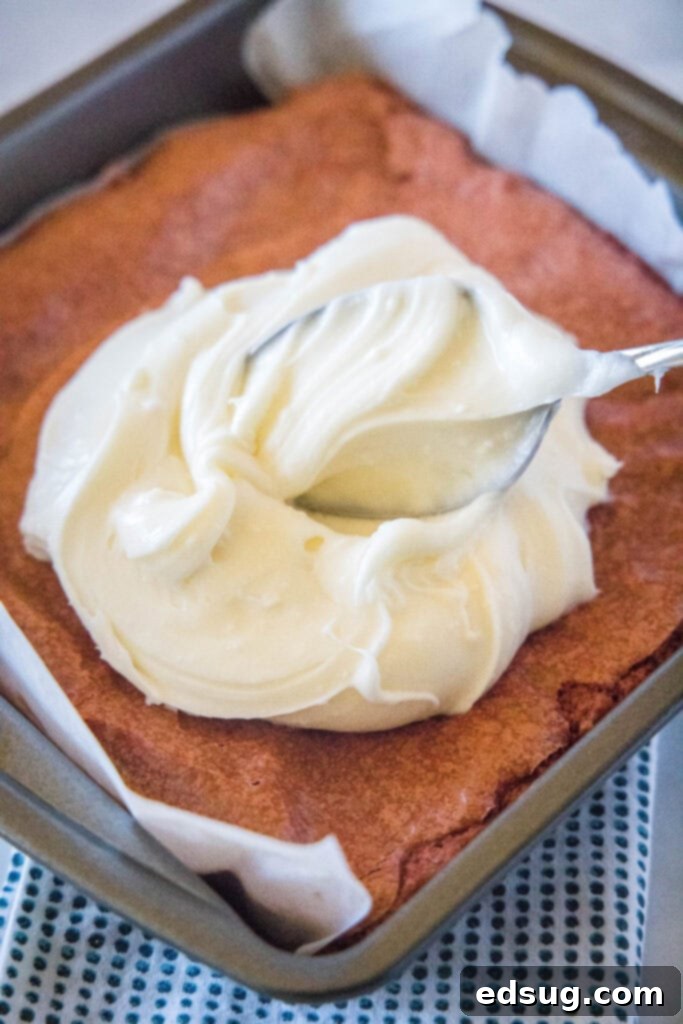 A hand spreading white cream cheese frosting evenly over a pan of cooled red velvet brownies.