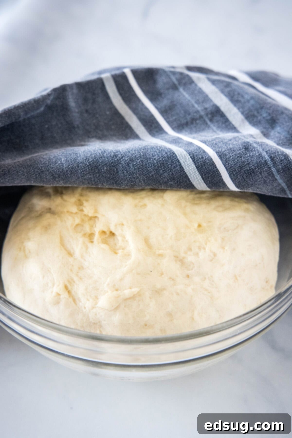 Risen bread dough, visibly doubled in size, covered with a dishcloth in a clear glass bowl, indicating successful proofing.