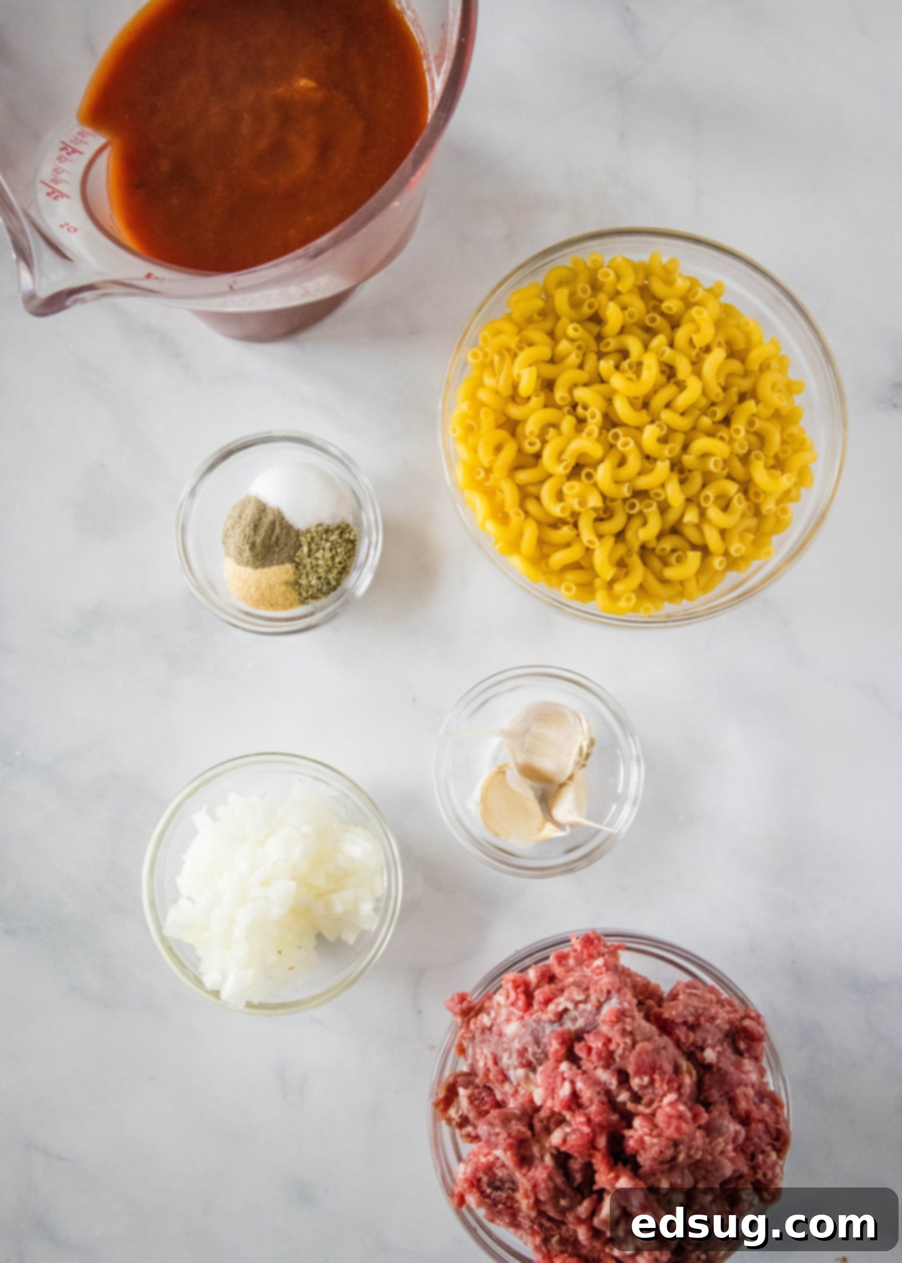 Overhead view of all the fresh ingredients needed for beefaroni: a bowl of macaroni, a bowl of lean ground beef, a pitcher of rich tomato sauce, a small bowl of mixed seasonings, a bowl of minced garlic, and a bowl of chopped onions, all arranged on a rustic surface.