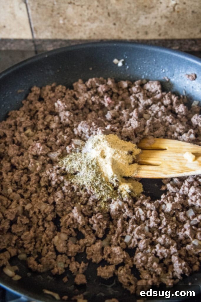 Ground beef cooking in a large skillet, with chopped onions and minced garlic, along with Italian seasonings scattered on top, ready to be mixed to form the base of the beefaroni sauce. A wooden spoon rests beside it.