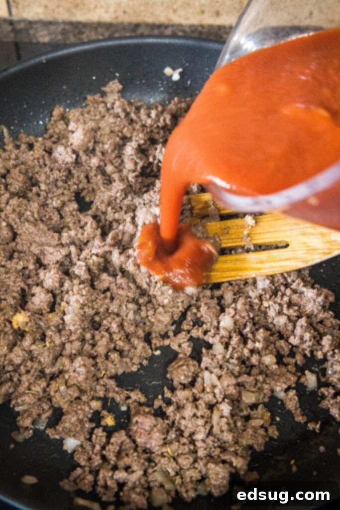 Thick tomato sauce being poured from a jar into a skillet containing already browned and seasoned ground beef and onions, beginning to form the hearty beefaroni sauce.