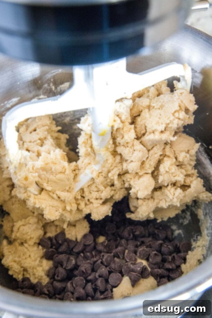A stand mixer paddle mixing cookie dough with a pile of semisweet chocolate chips being incorporated.