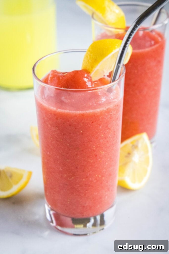 A tall glass brimming with vibrant pink frozen strawberry lemonade, garnished with a fresh lemon slice and a strawberry.