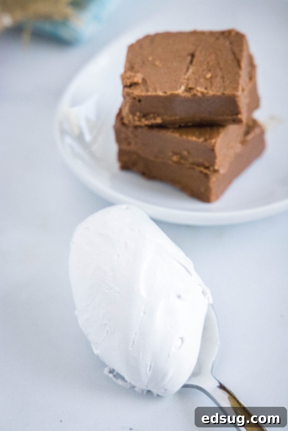 This easy homemade marshmallow fluff recipe is creamy, pillowy, packed with vanilla flavor, and so much more tasty than the store-bought kind. A spoonful of homemade marshmallow fluff next to a plate with two pieces of fudge, highlighting its creamy texture.