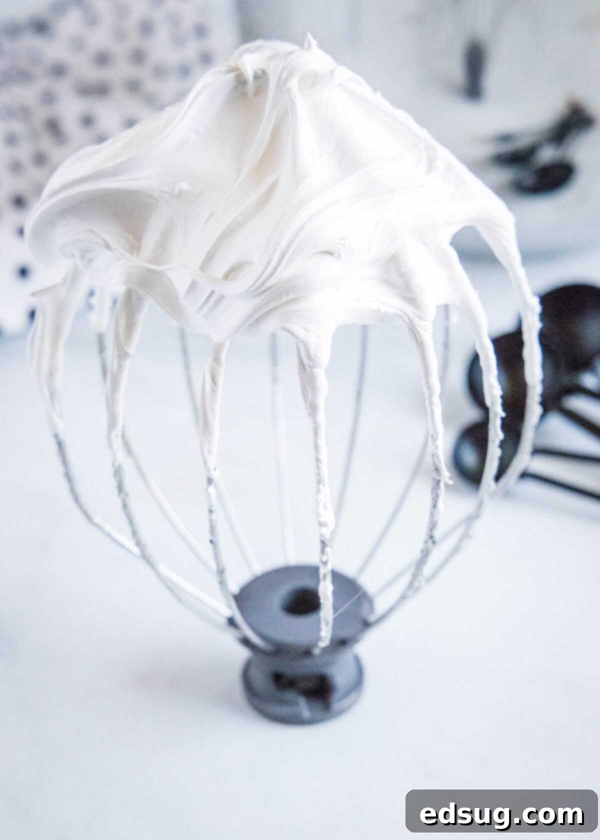 This easy homemade marshmallow fluff recipe is creamy, pillowy, packed with vanilla flavor, and so much more tasty than the store-bought kind. Close up of a stand mixer whisk attachment covered in stiff, perfectly formed peaks of marshmallow fluff, showcasing its ideal texture.
