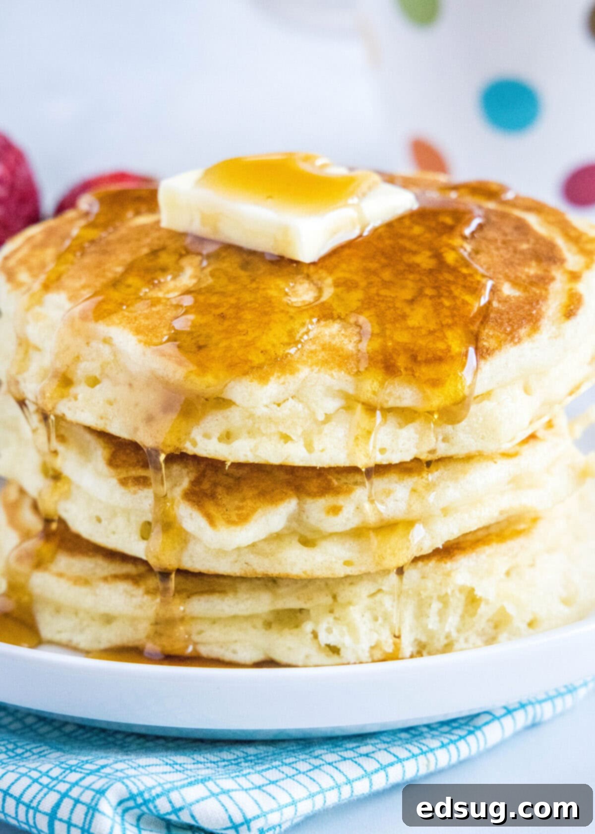 Close up of a tall stack of golden-brown buttermilk pancakes, generously topped with a pat of melting butter and glistening maple syrup cascading down the sides. The pancakes are thick and look incredibly soft.