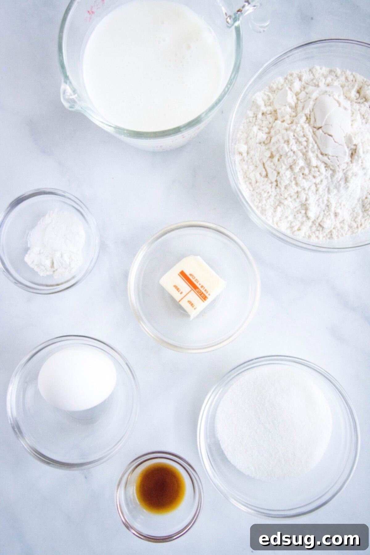 An overhead view of all the essential ingredients laid out on a clean surface, ready for making buttermilk pancakes. Includes a bowl of all-purpose flour, a glass measuring cup filled with buttermilk, a bowl of granulated sugar, a small dish of vanilla extract, a bowl containing baking powder and baking soda, a stick of butter, and a single egg.