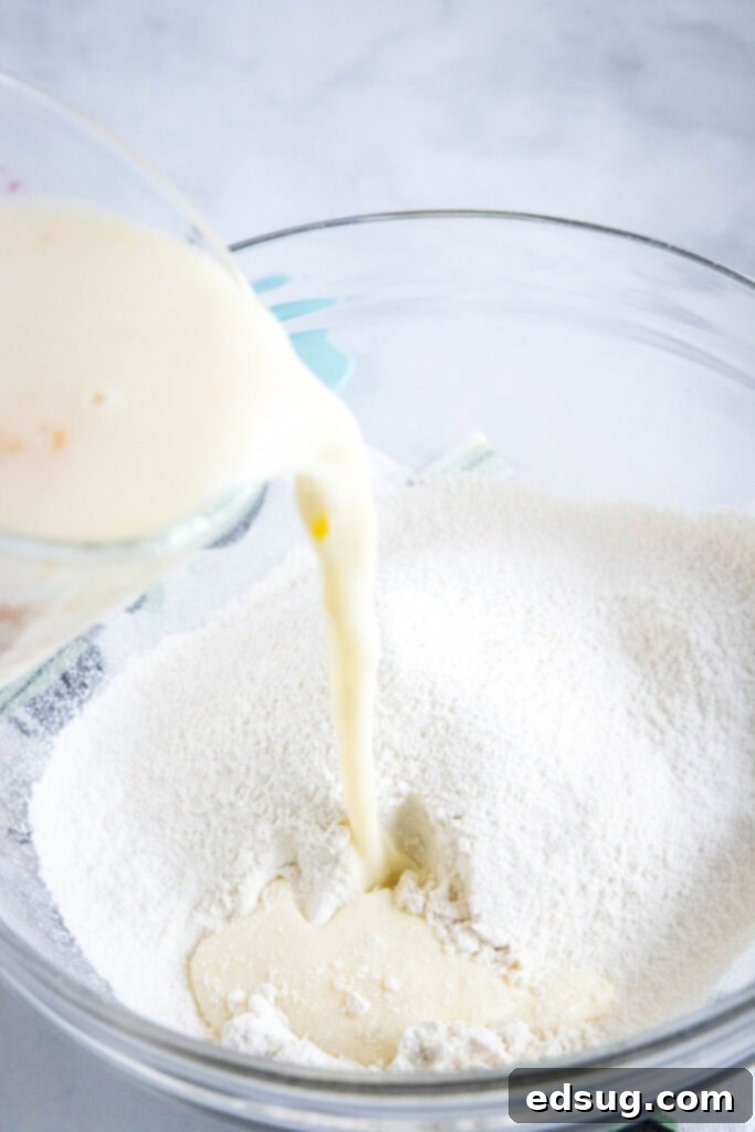 A hand pouring a smooth liquid buttermilk mixture from a Pyrex measuring cup into a large bowl of dry flour mixture, preparing the pancake batter.