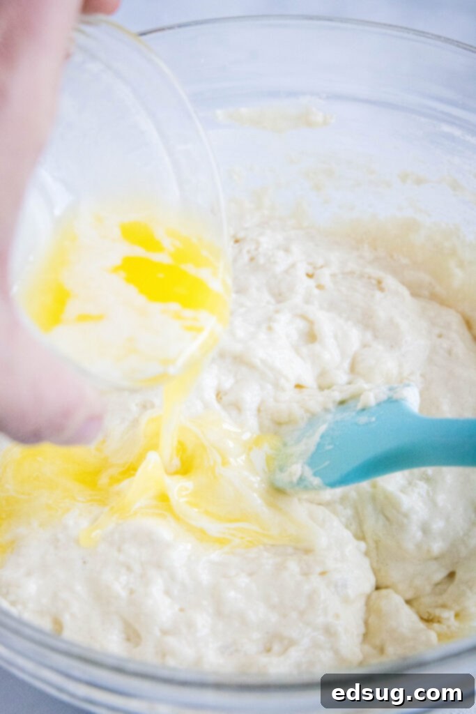 A hand gently pouring melted butter into a bowl of creamy pancake batter, with a spatula resting nearby, highlighting the final step of batter preparation.