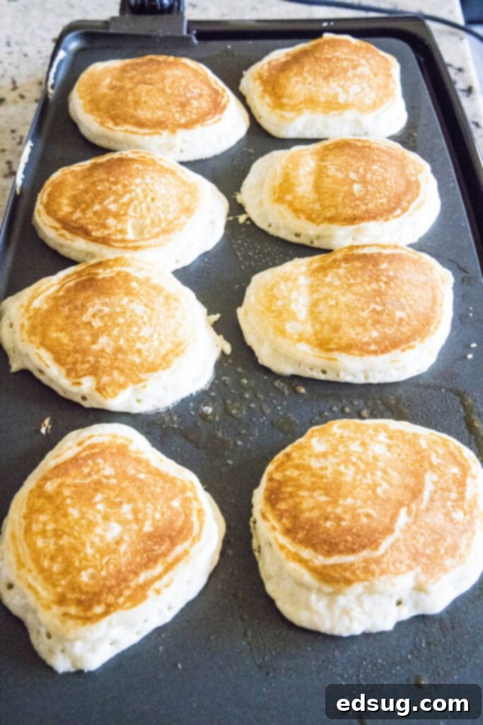 Eight perfectly round, golden-brown buttermilk pancakes sizzling on a large electric griddle, with some just flipped and others still cooking, showcasing a busy breakfast scene.