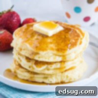 A stack of golden-brown buttermilk pancakes on a white plate, garnished with a pat of butter, a drizzle of syrup, and fresh strawberries and blueberries in the background.