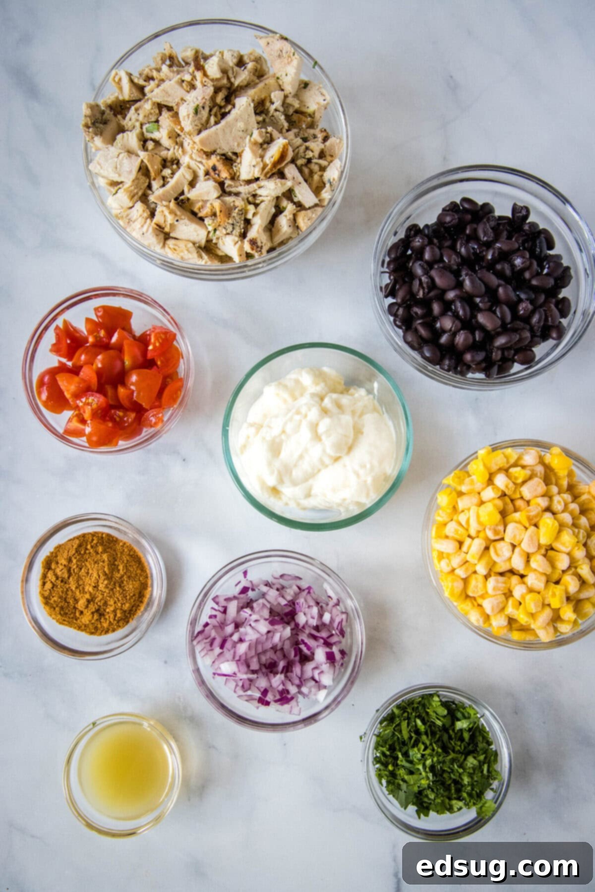 Overhead view of the ingredients needed for southwest chicken salad: a bowl of shredded chicken, a bowl of black beans, a bowl of chopped tomatoes, a bowl of mayo, a bowl of corn, a bowl of chopped onion, a bowl of cilantro, a bowl of taco seasoning, and a bowl of lime juice
