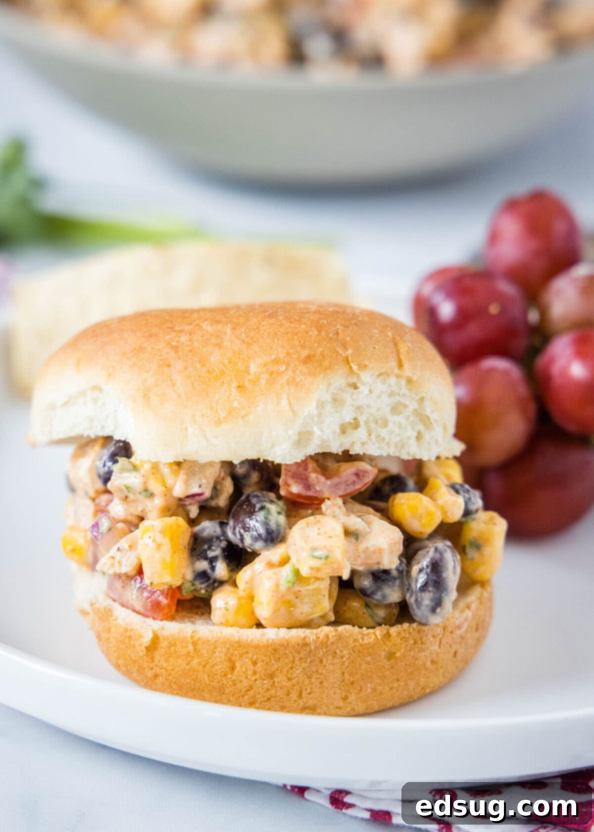 A burger bun filled with chicken salad with corn and beans, on a plate next to some grapes