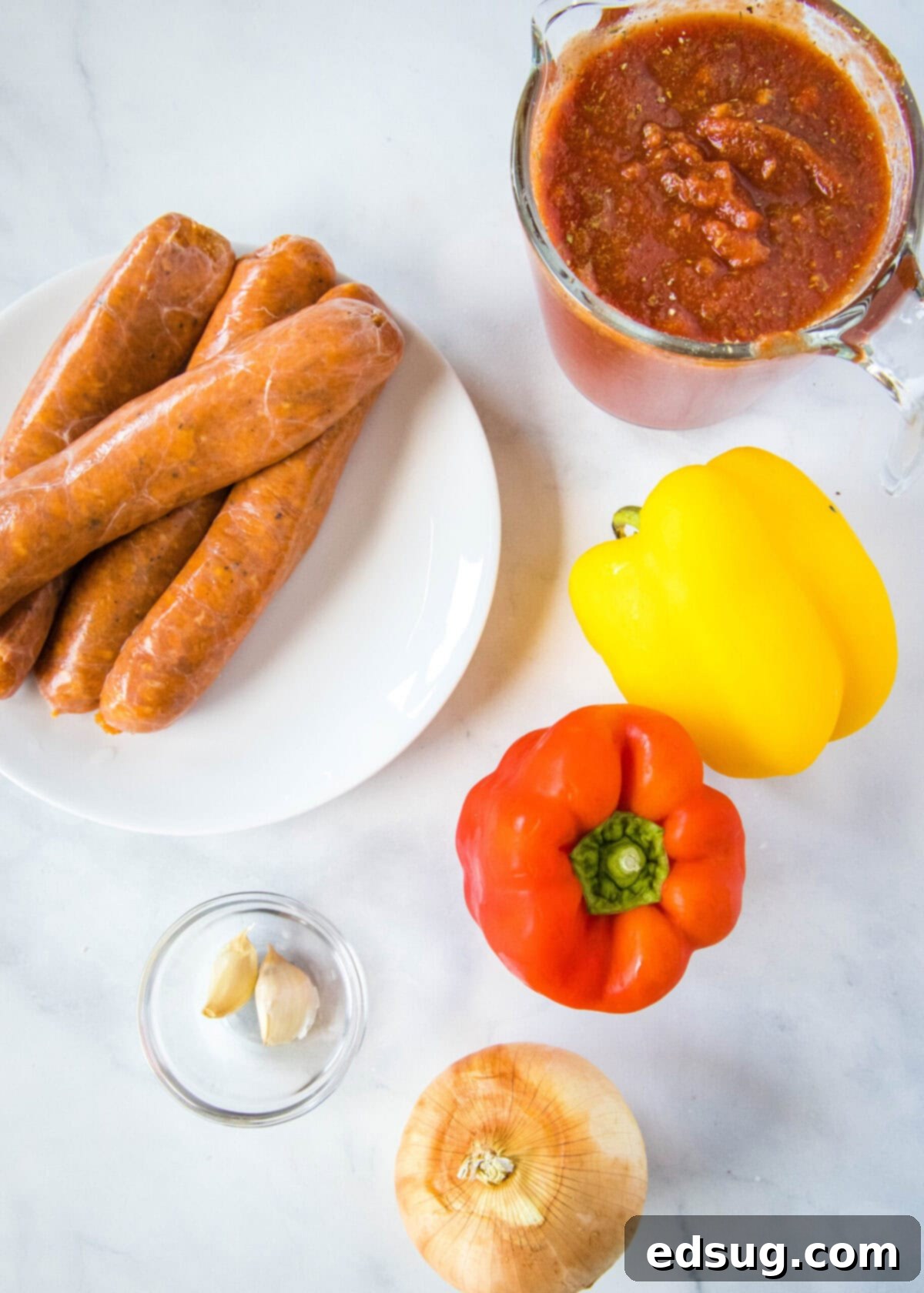 Overhead view of the ingredients needed for crock pot sausage and peppers: a plate of chicken sausage, a jar of marinara sauce, a yellow bell pepper, a red bell pepper, a yellow onion, and some garlic cloves