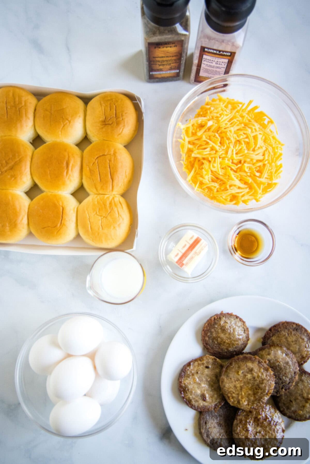 Overhead view of various ingredients for breakfast sliders: slider buns, sausage patties, eggs, milk, shredded cheddar cheese, butter, maple syrup, and seasonings.