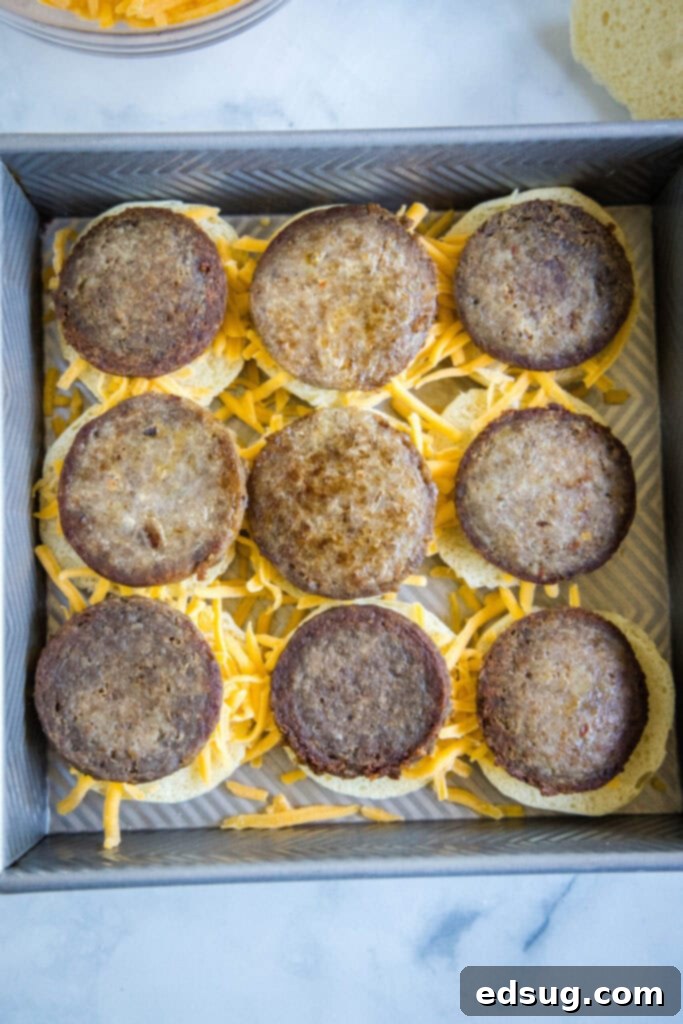 Nine sausage patties placed on a layer of shredded cheese atop slider buns in a baking dish.