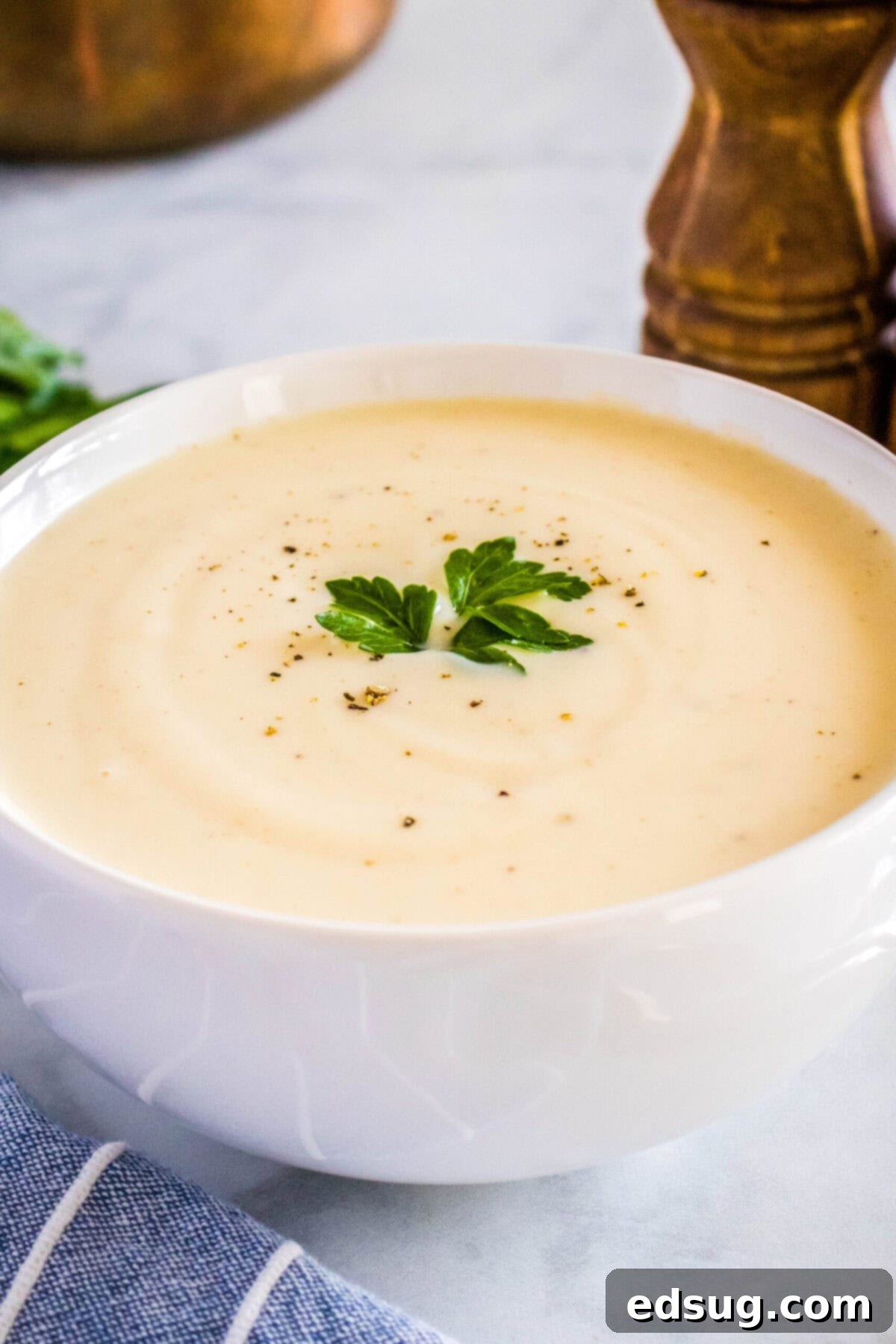 Close-up of a steaming bowl of homemade cream of chicken soup, garnished with fresh parsley and black pepper, with a pepper grinder in the background.