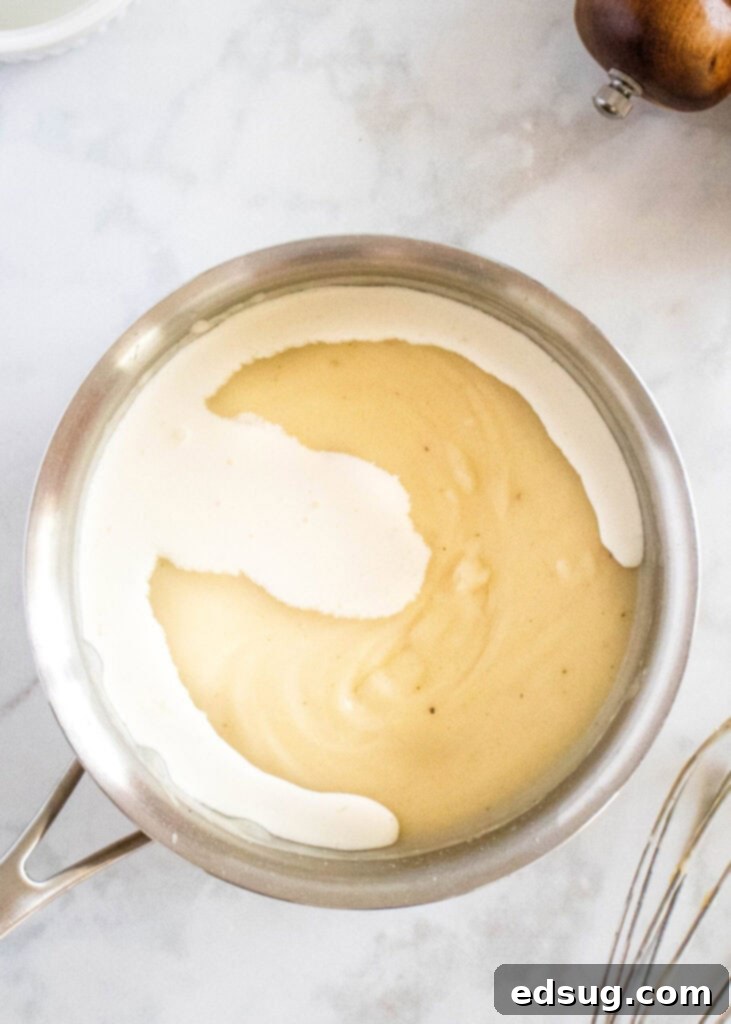 Overhead view of a saucepan filled with thickened cream of chicken soup base, with a swirl of rich heavy cream freshly poured on top, ready to be stirred in.