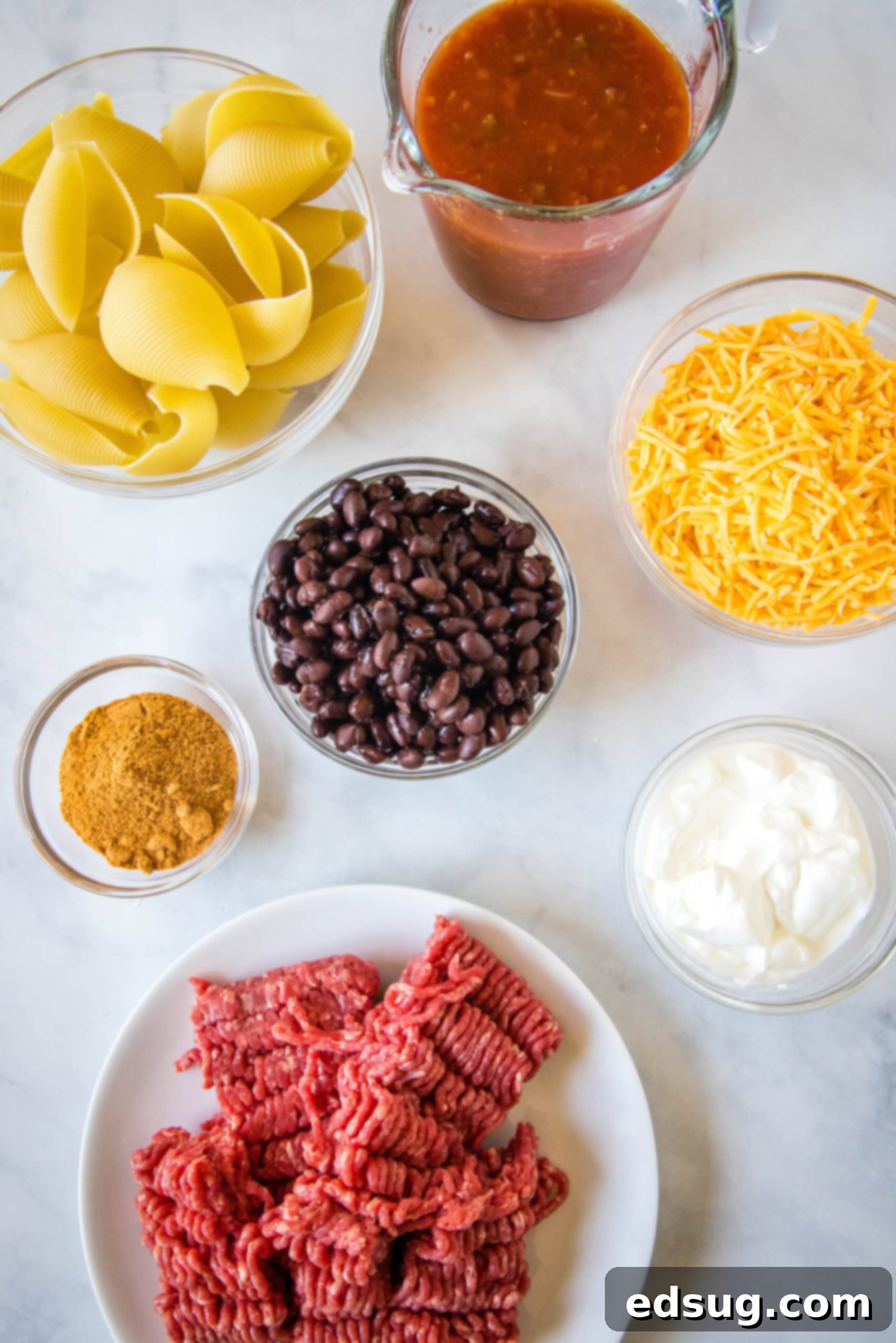 Various fresh ingredients laid out for making taco stuffed shells: jumbo pasta shells, ground beef, taco seasoning, black beans, salsa, shredded cheese, and sour cream.