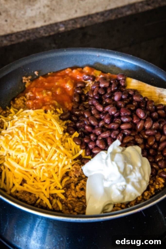 A large skillet containing browned taco meat, with salsa, black beans, sour cream, and shredded cheese being added to it, ready to be mixed into the creamy filling.