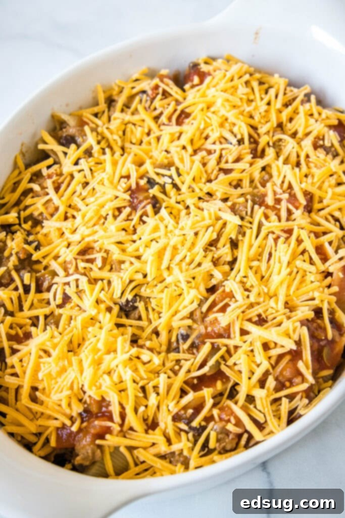 Assembled taco stuffed shells in a baking dish, now covered with both salsa and a generous layer of shredded cheese, perfectly prepped for baking.