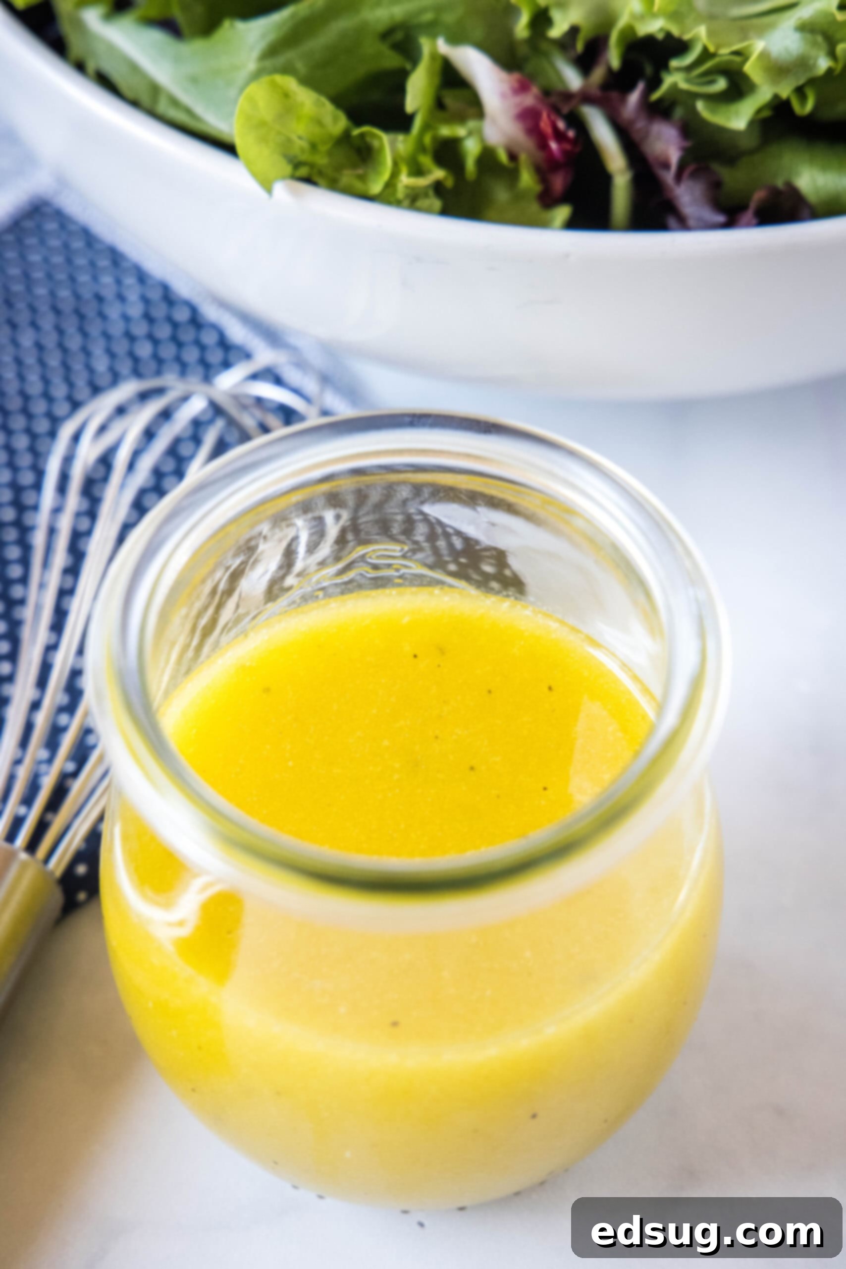 A jar of apple cider vinaigrette on a countertop next to a whisk and a bowl of salad.