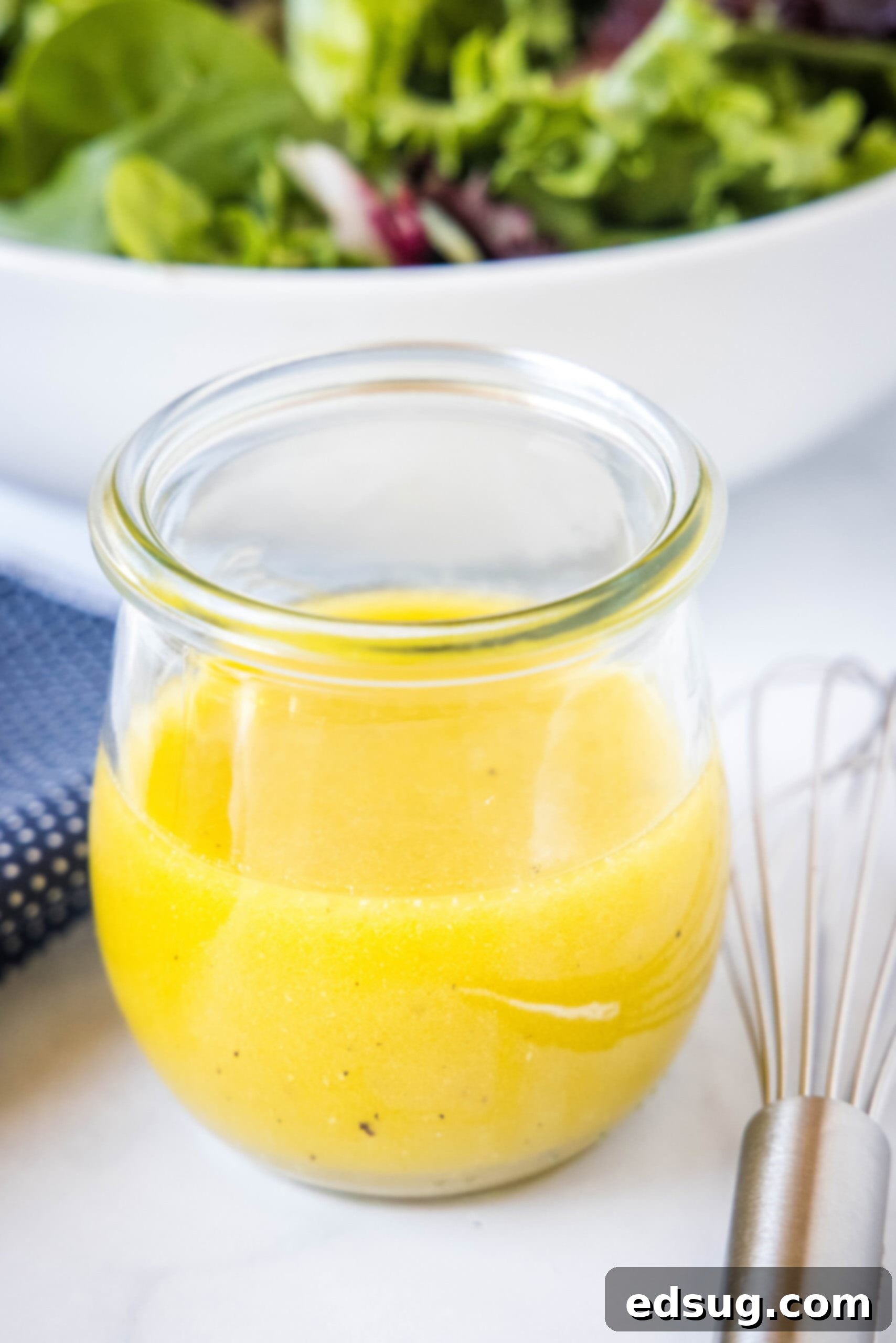 A jar of apple cider vinaigrette on a countertop next to a whisk and a bowl of salad.
