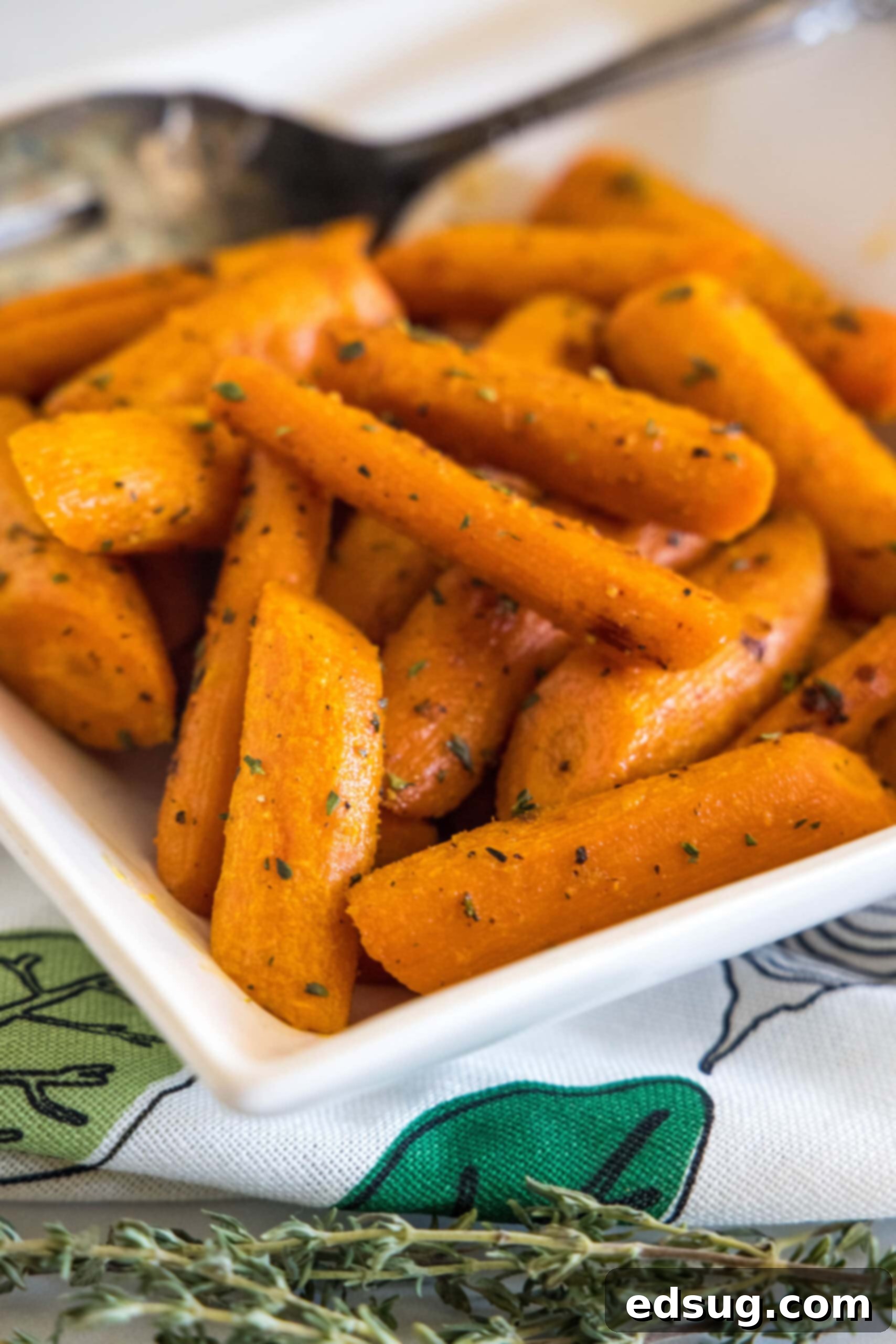 Seasoned roasted carrots in a square white bowl with a serving spoon, garnished with fresh herbs, close up.