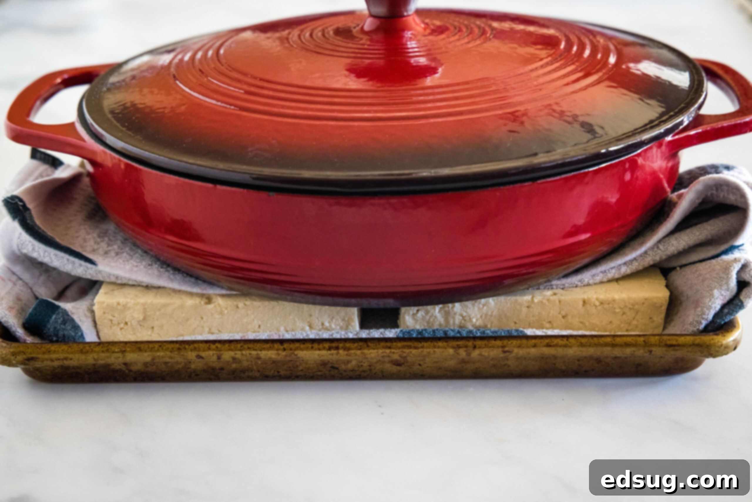 A large red skillet placed on top of a block of tofu wrapped in clean kitchen towels, demonstrating the tofu pressing technique.