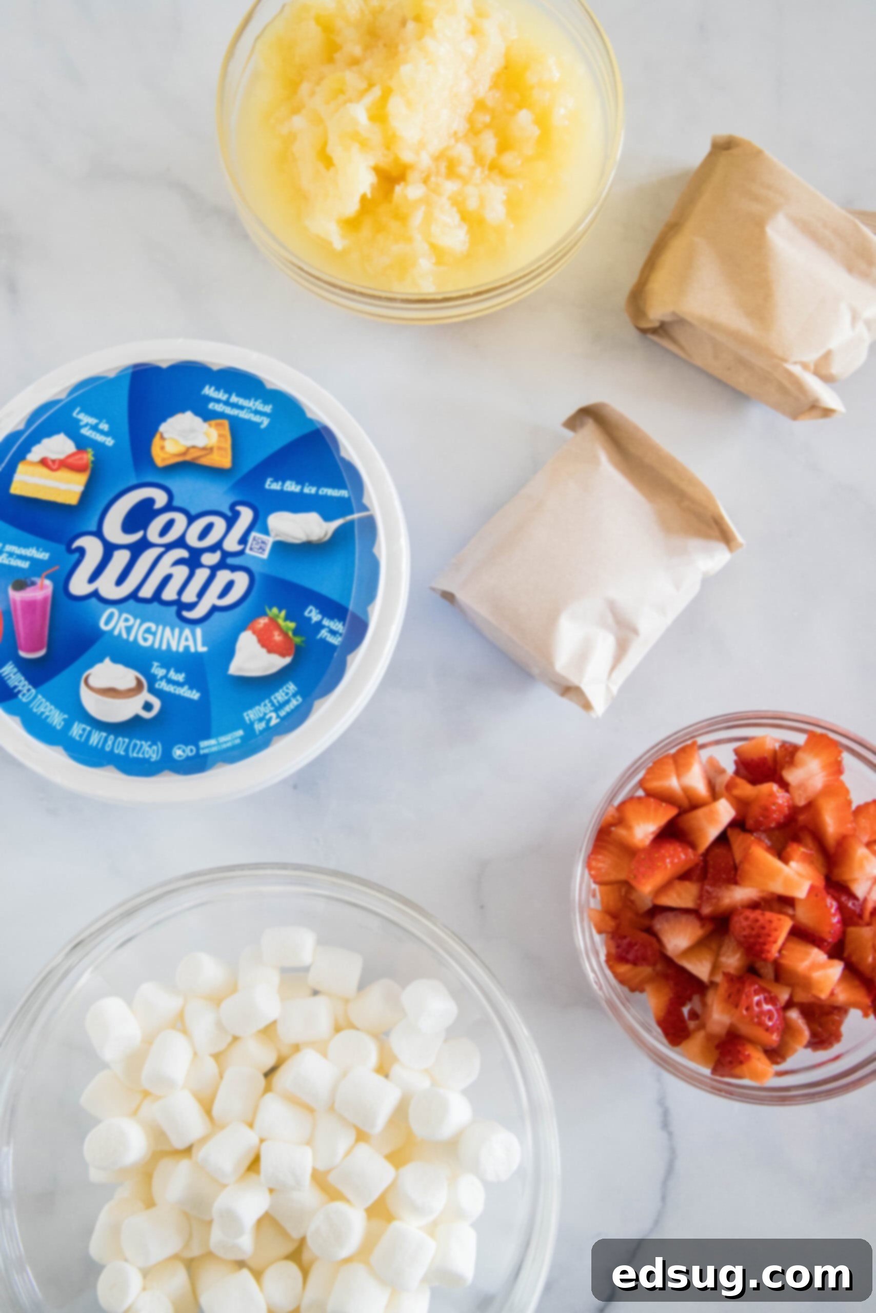 Ingredients for strawberry fluff salad laid out on a table, including Jello mix, pudding mix, Cool Whip, strawberries, pineapple, and marshmallows.
