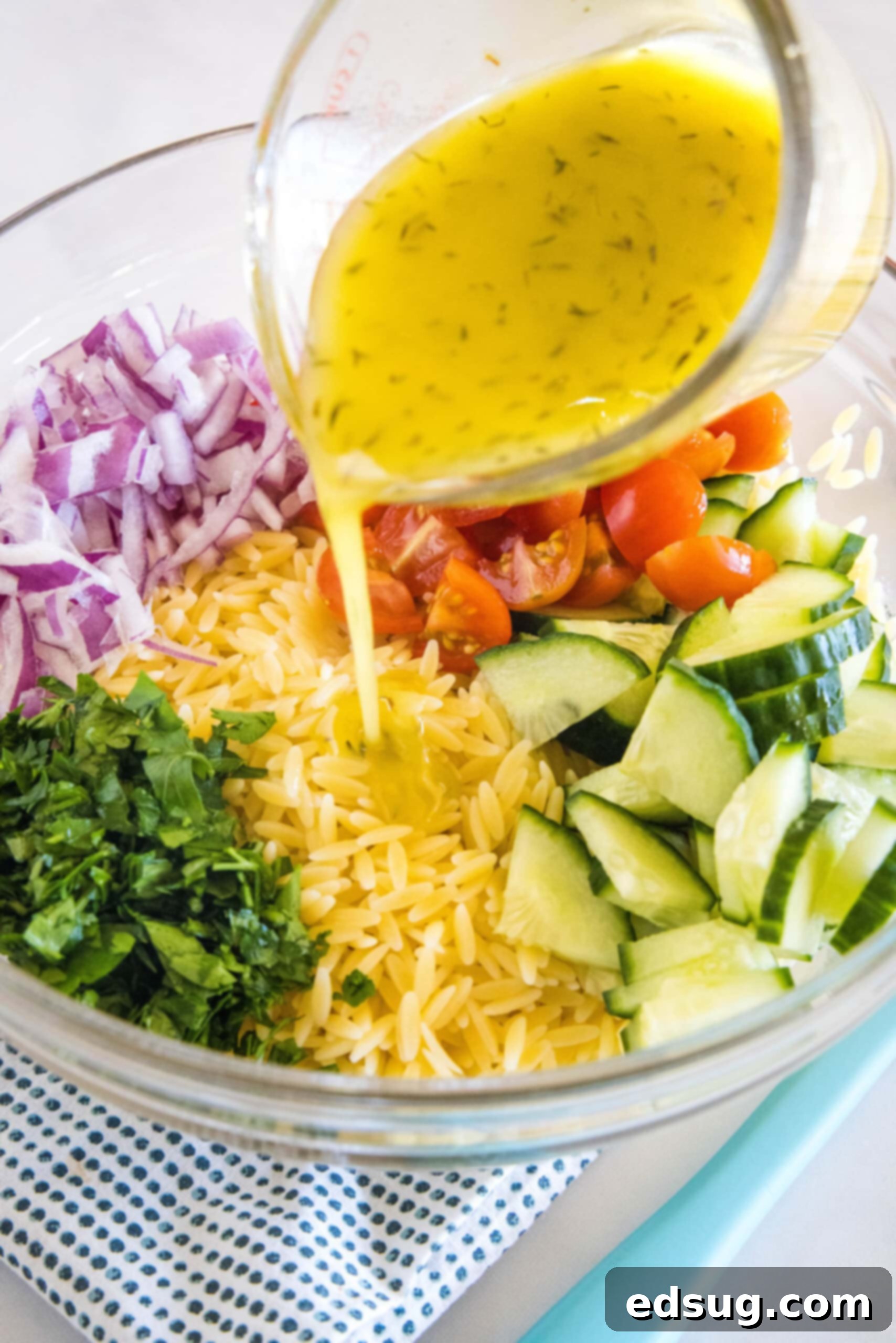 Lemon vinaigrette being poured from a measuring cup over orzo pasta salad ingredients in a large glass bowl, illustrating the dressing step.