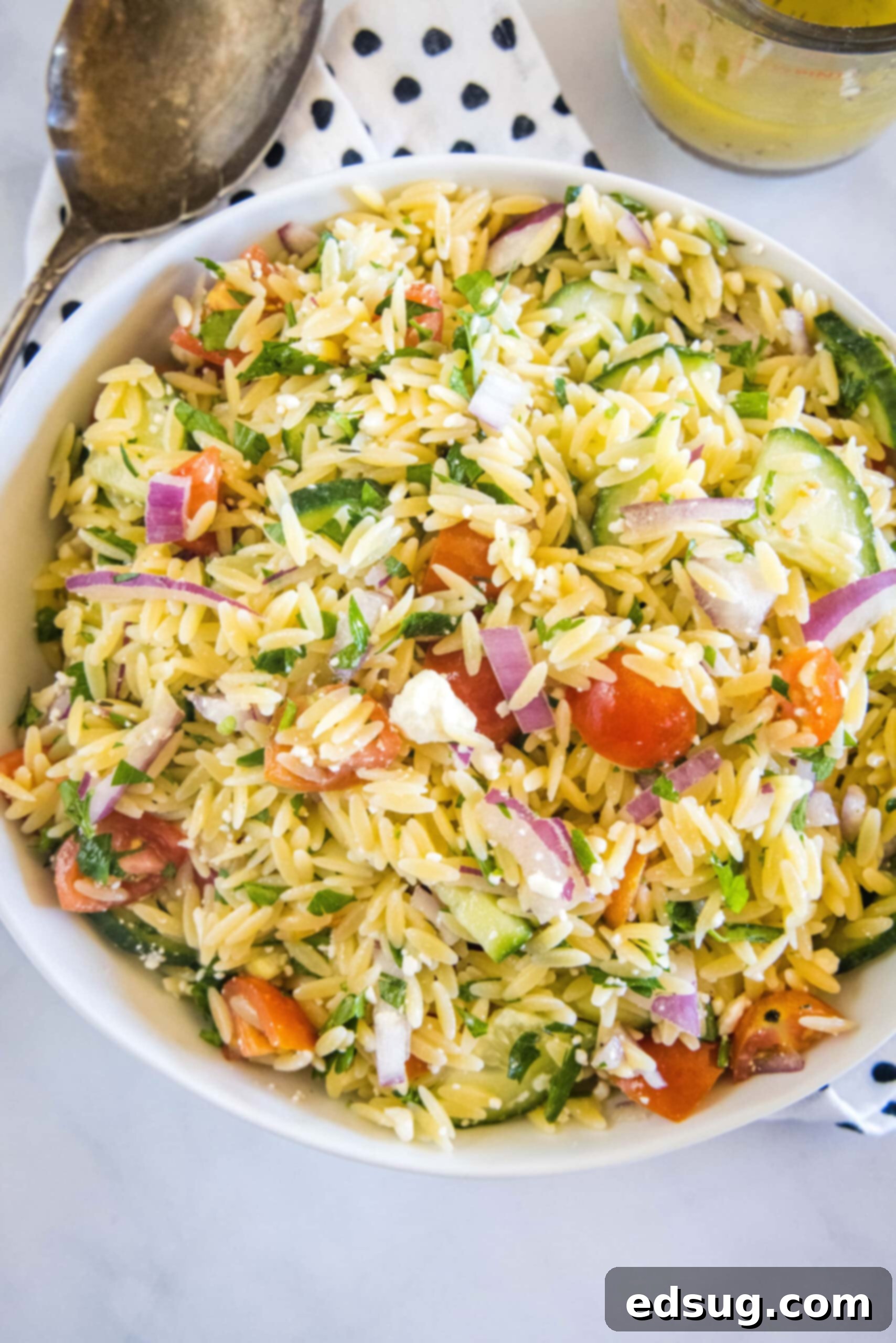 Overhead view of orzo pasta salad in a large bowl next to a serving spoon and a jar of dressing, showcasing the final dish.