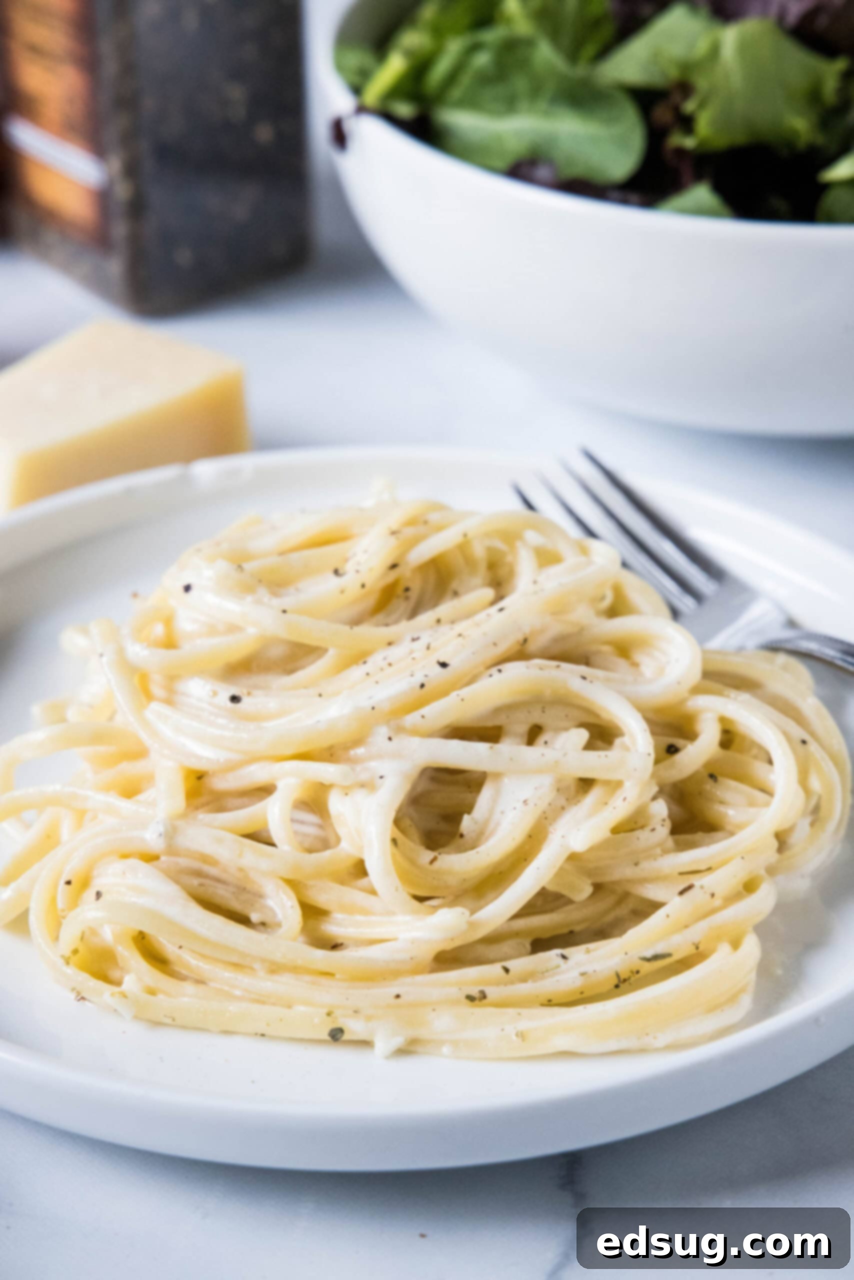 This easy homemade Alfredo sauce is rich and creamy and packed with garlicky parmesan flavors. Make it in one pan with your favorite pasta! A serving of pasta alfredo on a white plate next to a fork, with a bowl of salad in the background.