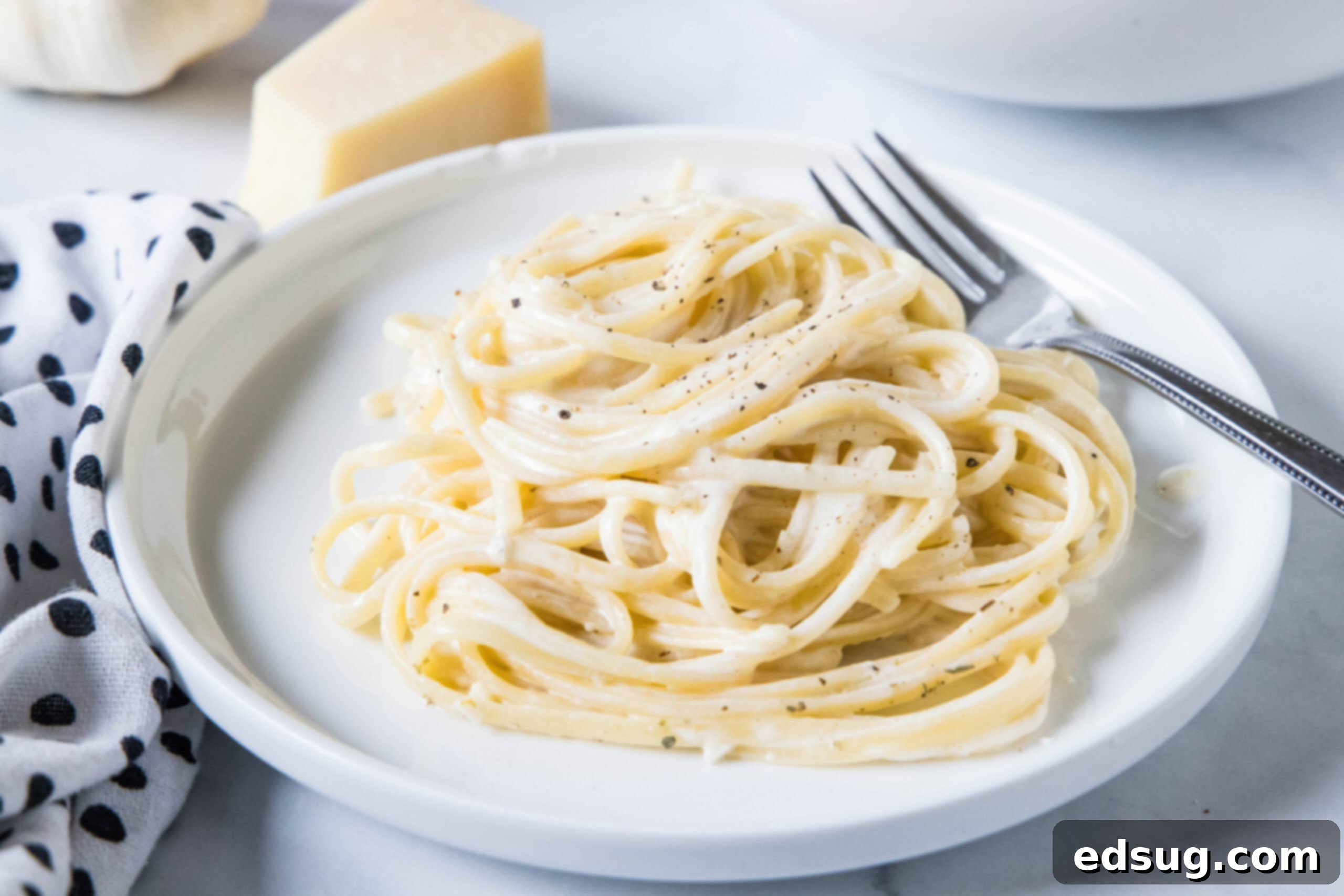 This easy homemade Alfredo sauce is rich and creamy and packed with garlicky parmesan flavors. Make it in one pan with your favorite pasta! A serving of pasta alfredo on a white plate next to a fork, with a wedge of parmesan in the background.