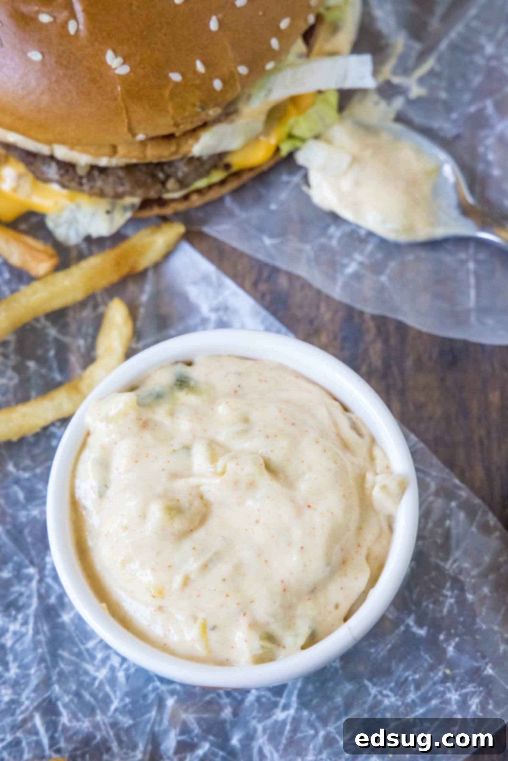 Overhead view of homemade Big Mac Sauce in a small serving bowl, glistening, next to a perfectly grilled burger and a side of crispy fries.