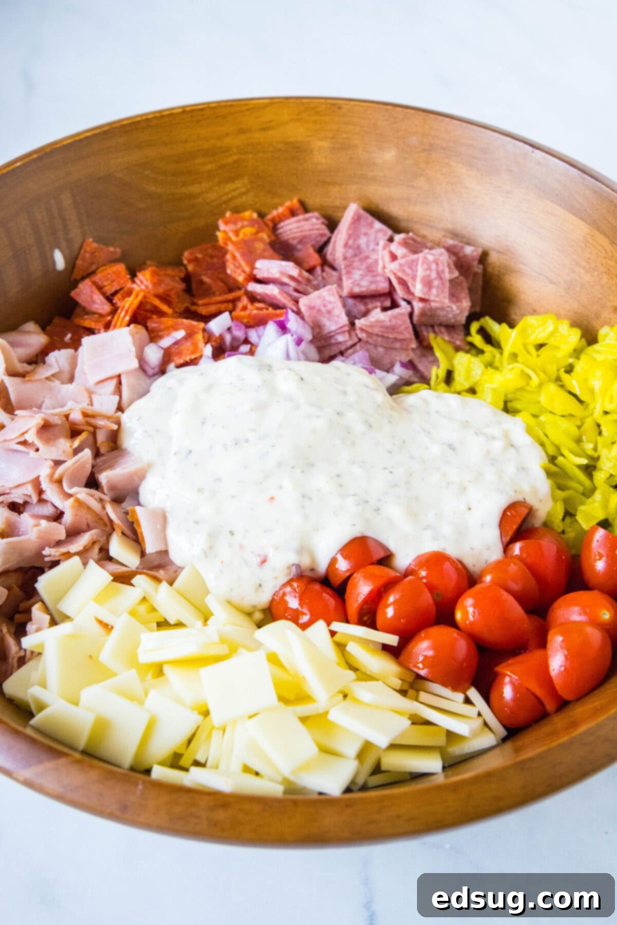 Pouring homemade creamy dressing over chopped deli meats, vegetables, and cheese in a large wooden salad bowl for hoagie dip.