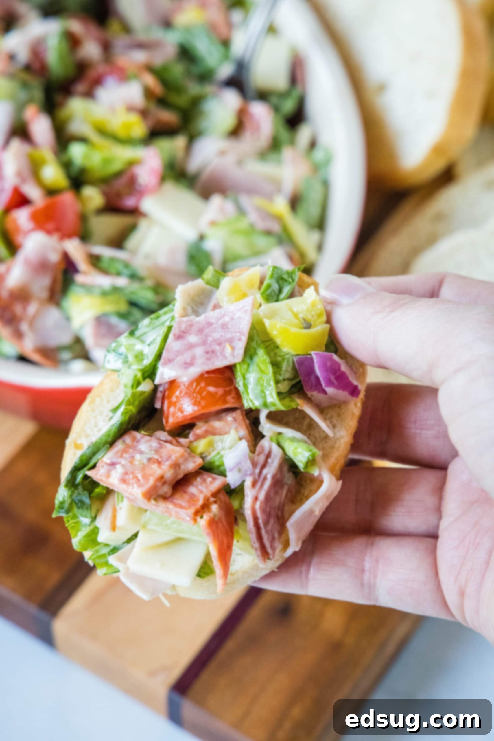A hand holds up a piece of crusty bread topped with a generous scoop of hoagie dip, highlighting its vibrant colors and creamy texture, with the full dip bowl and more bread in the background.