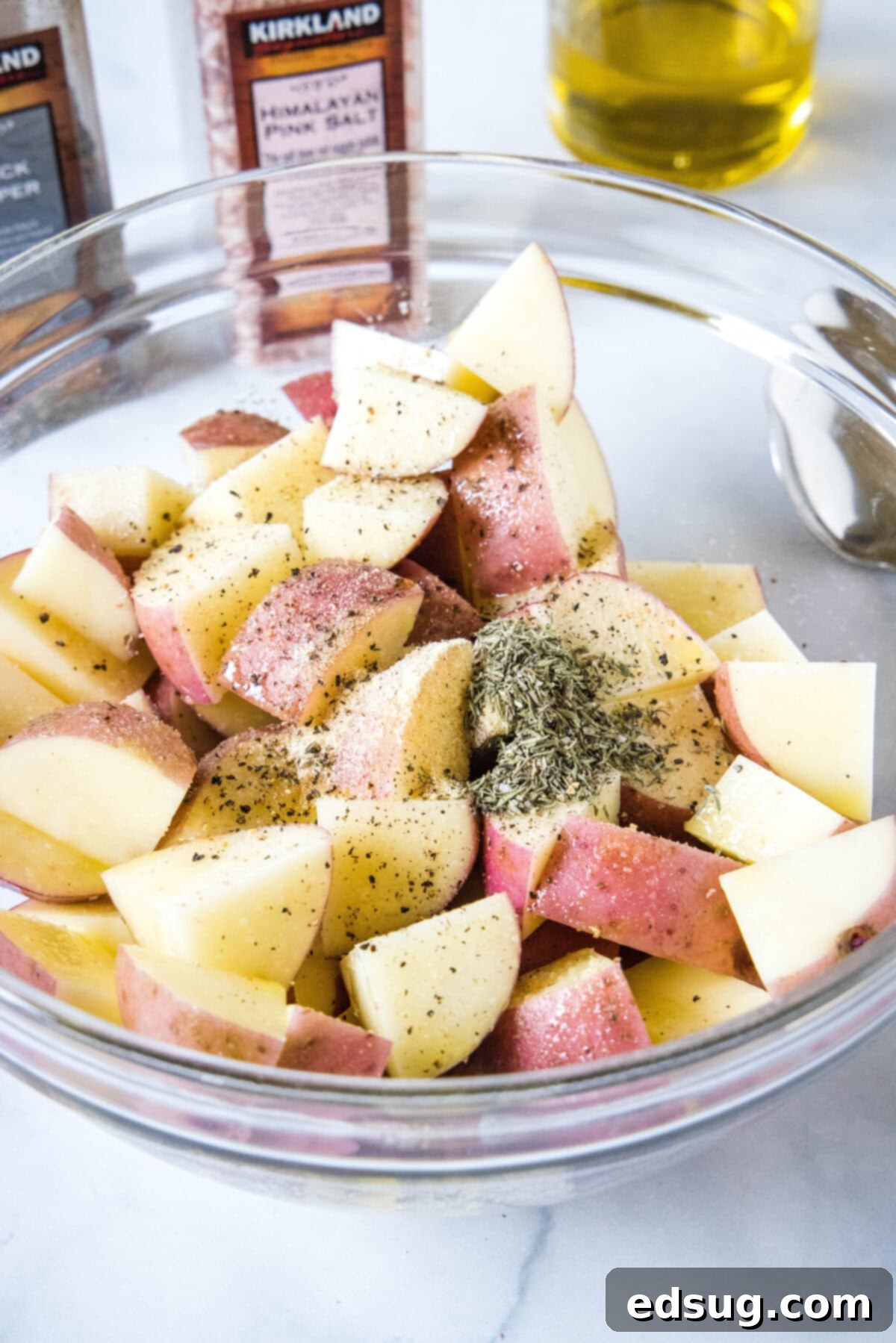 Seasonings being added to diced red potatoes in a glass mixing bowl.