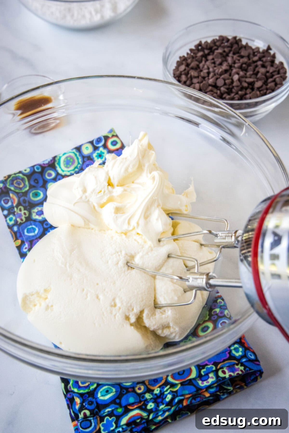 A hand mixer blending creamy ricotta and mascarpone cheese in a clear glass bowl, showcasing the smooth texture forming for the cannoli dip base.