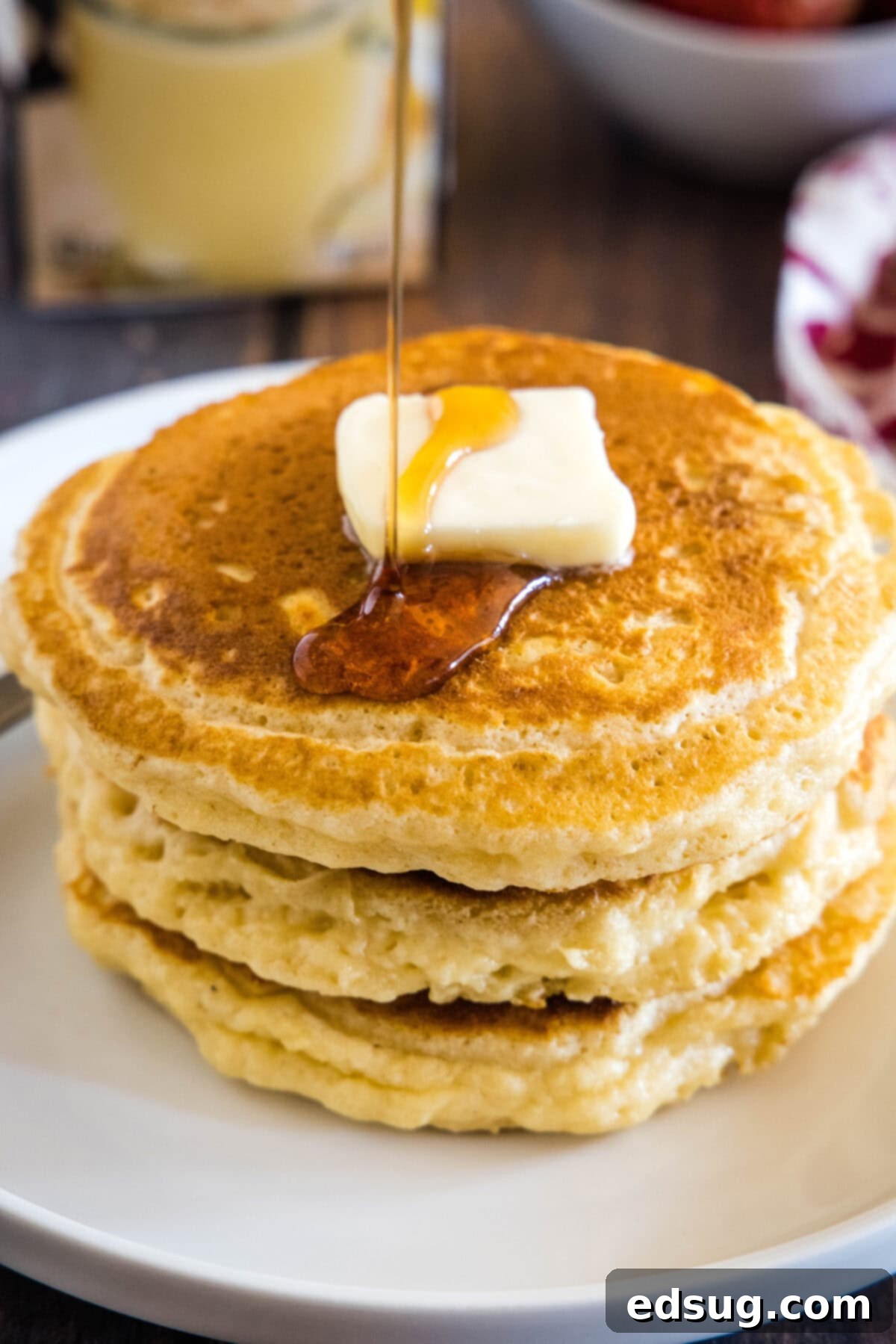 Festive Eggnog Pancakes 6 Maple syrup being poured over a stack of fluffy eggnog pancakes on a white plate, ready for breakfast.