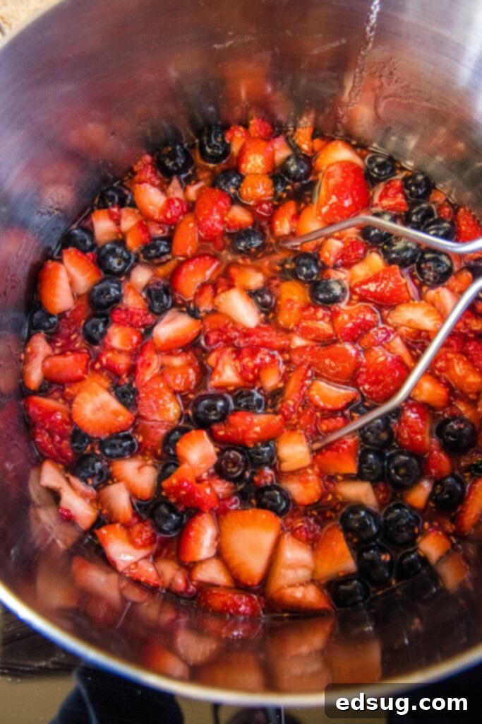 Berry Medley Jam 4 Macerated mixed berries in a stock pot with a potato masher resting inside, ready for cooking.