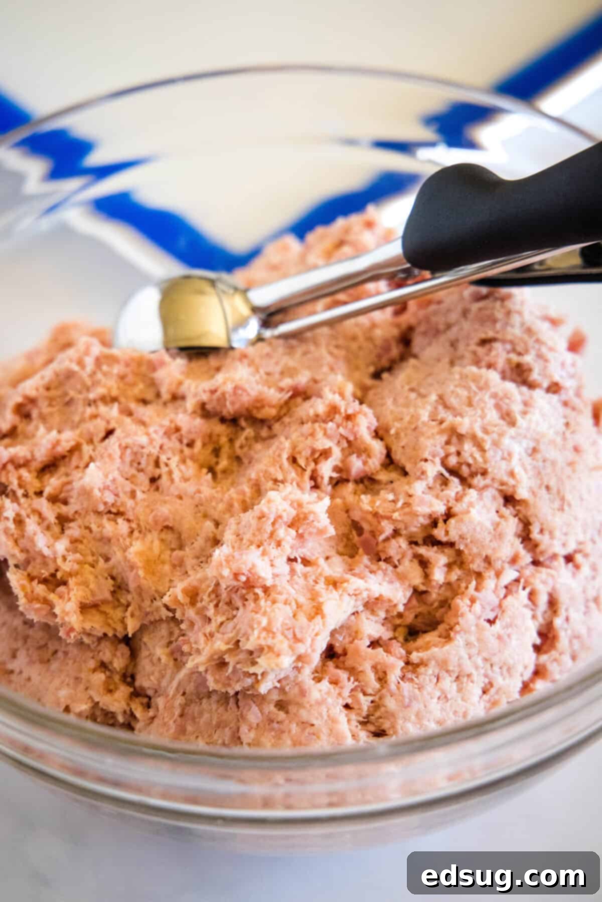 Portioning the ham ball mixture from a glass bowl into uniform small balls using a cookie scoop.