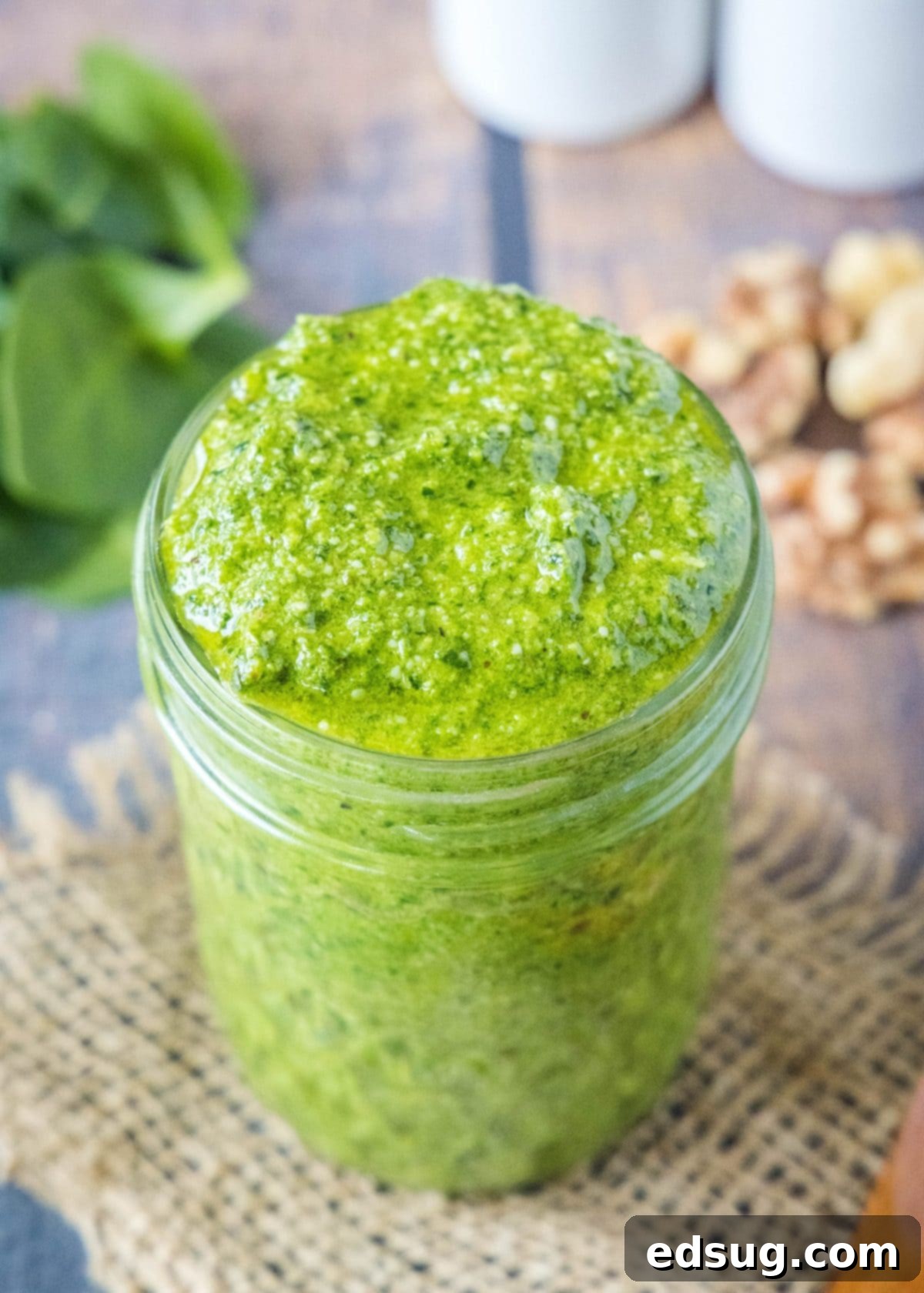 Close up of a jar of spinach pesto with fresh spinach and walnuts in the background, highlighting its natural ingredients.