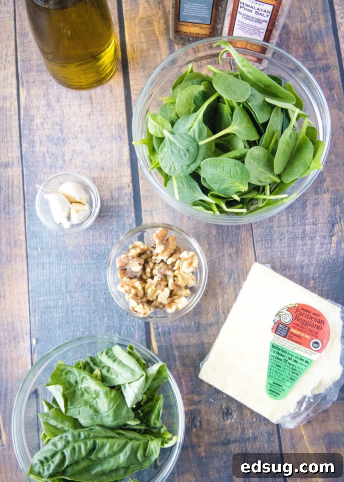 Overhead view of all the fresh ingredients needed for spinach pesto, including a vibrant bowl of spinach, aromatic basil, crunchy walnuts, peeled garlic cloves, a wedge of Parmesan cheese, olive oil, and seasonings.