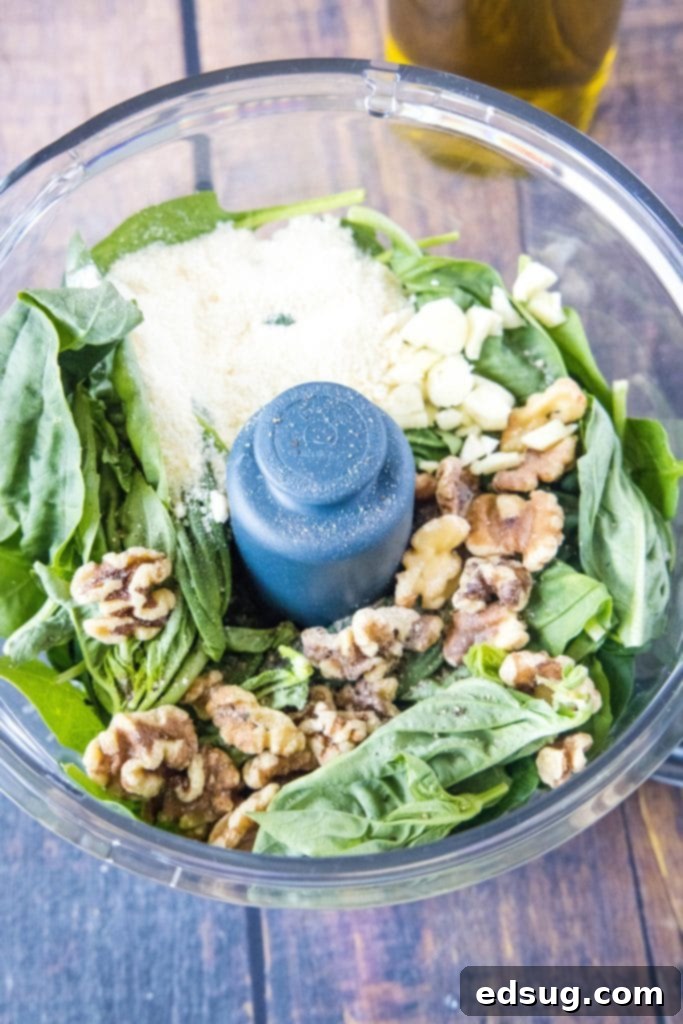 An overhead view of a food processor bowl filled with fresh garlic, grated Parmesan cheese, vibrant spinach, and aromatic basil leaves, ready for blending.