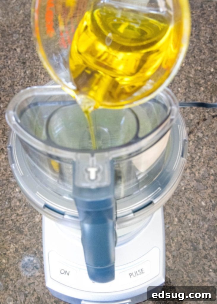 A Pyrex measuring cup pouring a steady stream of olive oil into a food processor while the ingredients are blending, to create a smooth emulsion.