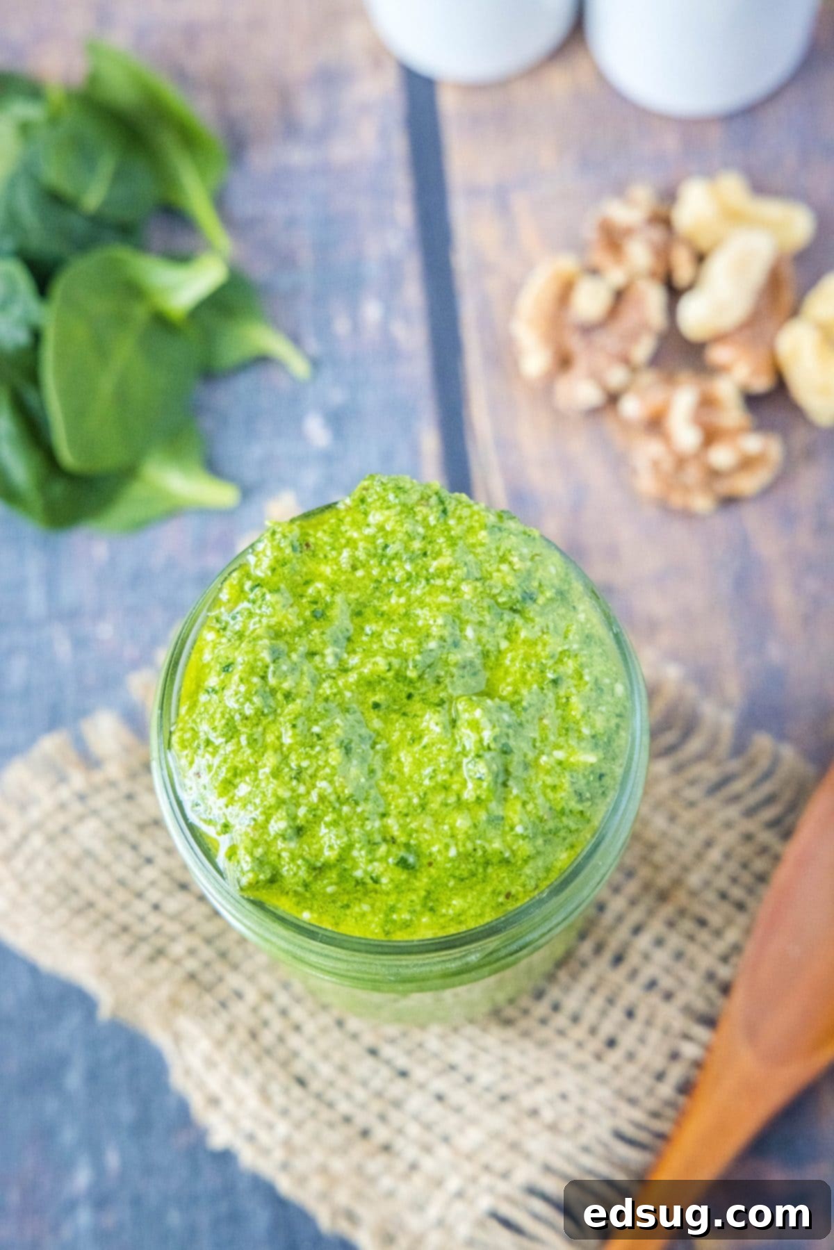 Overhead view of a jar of fresh spinach pesto, surrounded by a small pile of walnuts and fresh spinach leaves, showcasing its wholesome ingredients.
