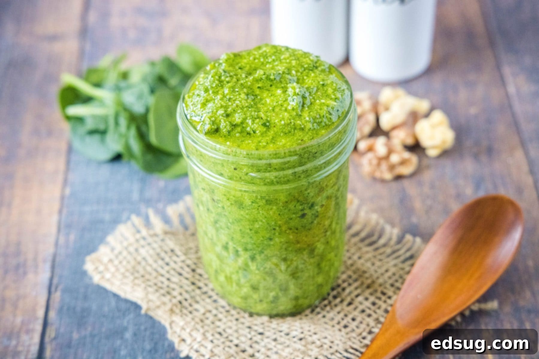 A jar of fresh spinach pesto prominently displayed, with whole walnuts and green spinach leaves artistically arranged in the background.